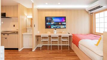 Habitación compacta con kitchenette, una televisión de pantalla plana montada en la pared, cama con manta roja y ventana con persianas.