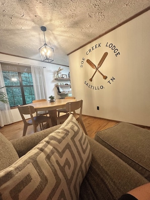 A cozy living room with a dining area, a wall-mounted sign reading 'Doe Creek Lodge Saltillo, TN', and a ceiling light fixture.