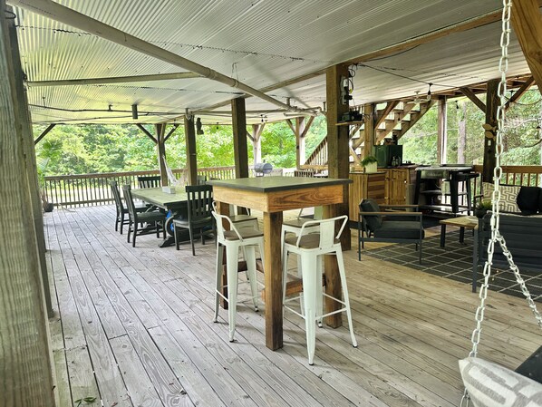 An outdoor covered patio area with a wooden deck, a dining table with chairs, a hanging swing, and a grill station.
