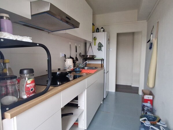 A kitchen with a stove, sink, and various kitchen utensils. There is a refrigerator and a window with curtains.