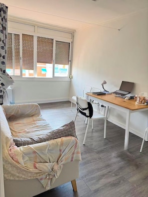 A room with a sofa, a wooden table with a laptop, a chair, and a window with blinds.