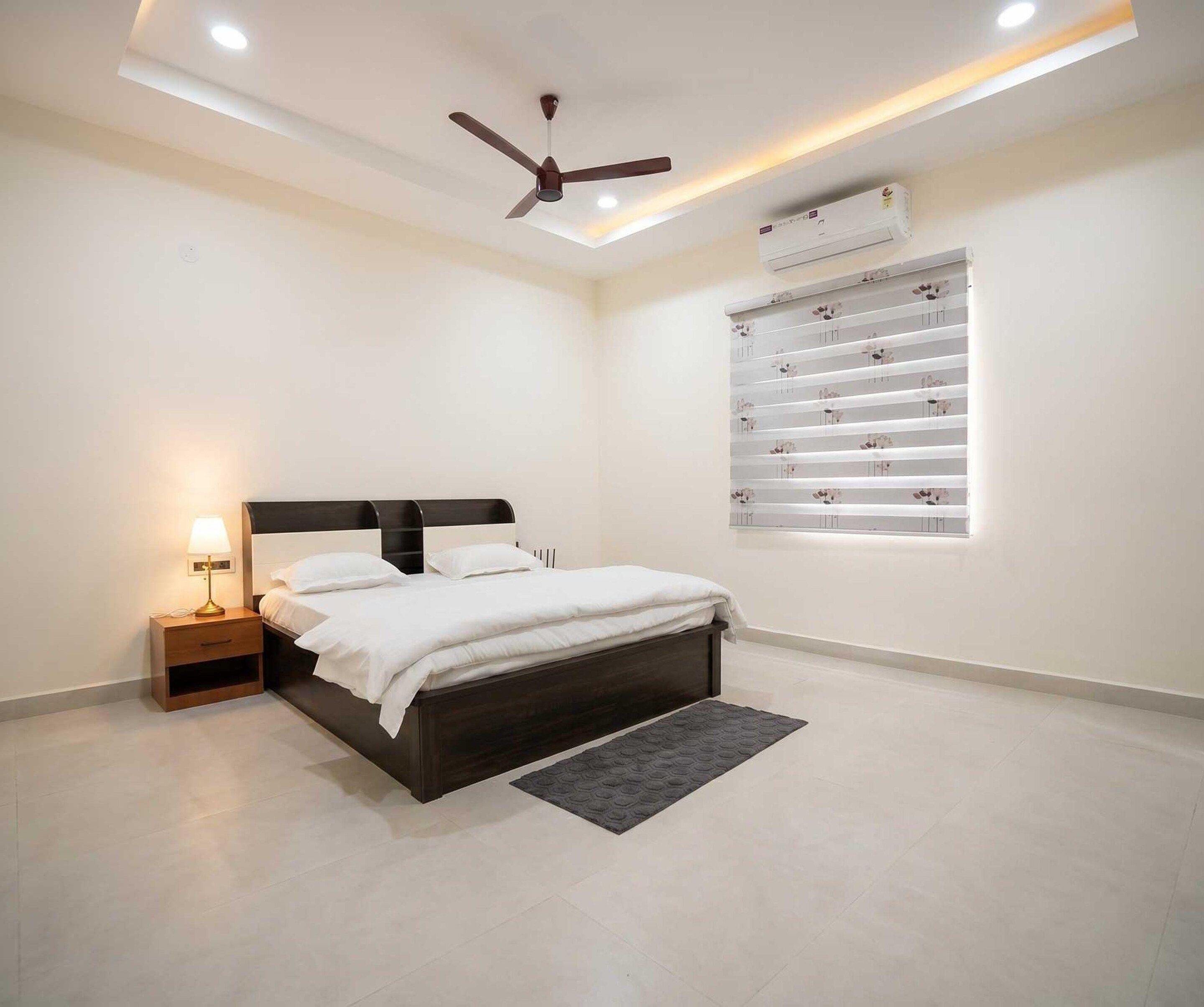 A bedroom with a bed, bedside table, lamp, ceiling fan, air conditioning unit, and a window with blinds.