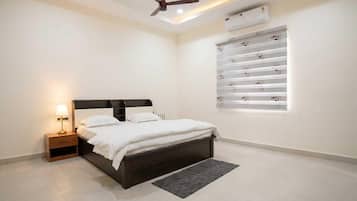 A bedroom with a bed, bedside table, lamp, ceiling fan, air conditioning unit, and a window with blinds.