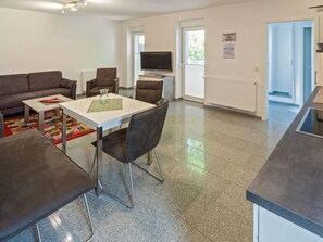 Ein modernes Wohnzimmer mit einer Couch, einem Couchtisch und einem Fernseher. Es gibt eine Küchenzeile mit einer Arbeitsplatte und einem Fenster.
