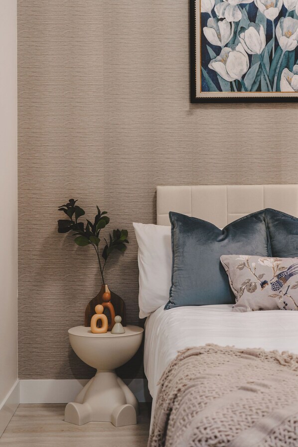 A bedroom with a bed, pillows, a bedside table with a plant, and a framed artwork on the wall.