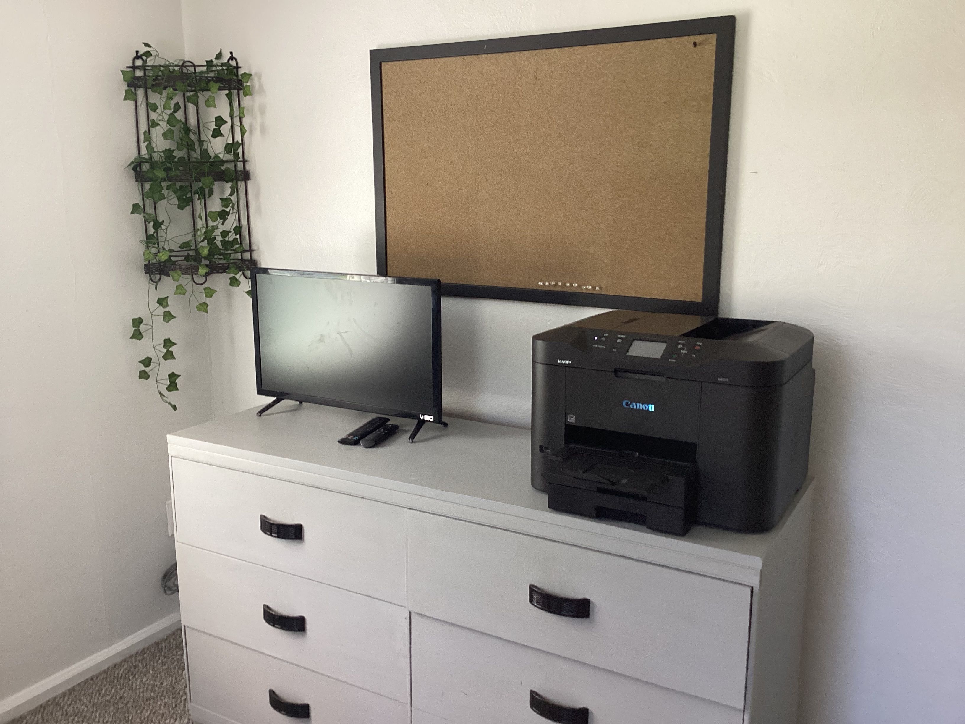 A white desk with a flat-screen TV, a printer, and a corkboard on the wall.
