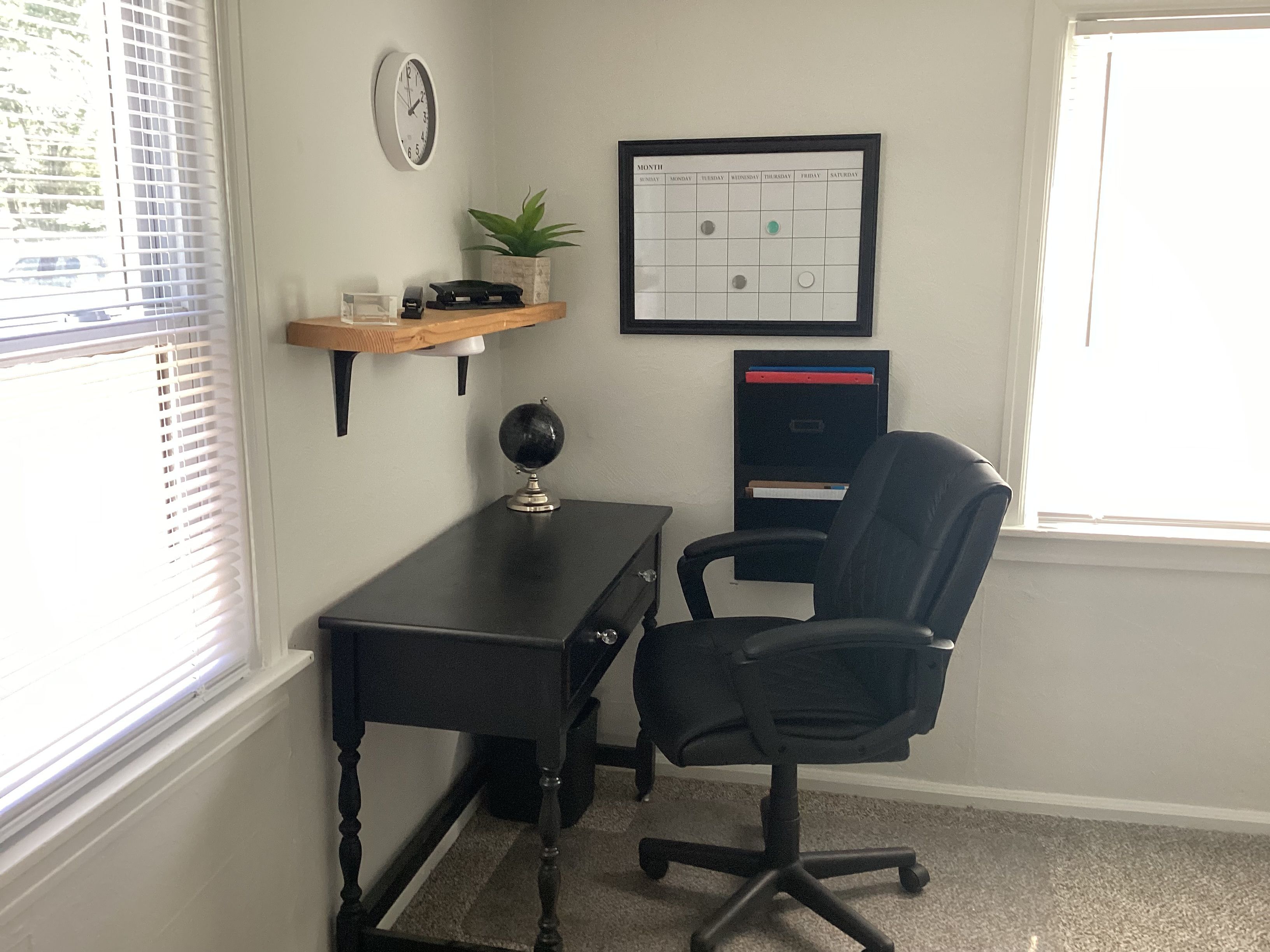 Une pièce comprenant un bureau, une chaise, une horloge, un calendrier et une fenêtre avec des stores.