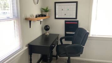 Une pièce comprenant un bureau, une chaise, une horloge, un calendrier et une fenêtre avec des stores.