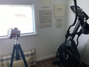 Quarto com bicicleta ergométrica, tripé com câmera e frases motivacionais na parede.