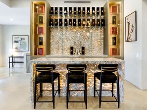 Uma área de bar com bancada de mármore, três banquetas pretas e prateleiras com diversos objetos.