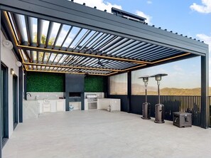 Uma área de cozinha externa com pergolado, parede verde e vista para as colinas.