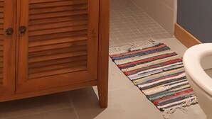 Ein Badezimmer mit einem Holzbadschrank, einem runden Spiegel, einem weißen Waschbecken, einem schwarzen Seifenspender, einem Streifen Teppich und einer weißen Toilette.