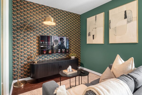 Un salon doté d’un téléviseur à écran plat fixé sur un mur orné de motifs, d’une console sombre, d’une table basse ronde et d’un canapé agrémenté de coussins.
