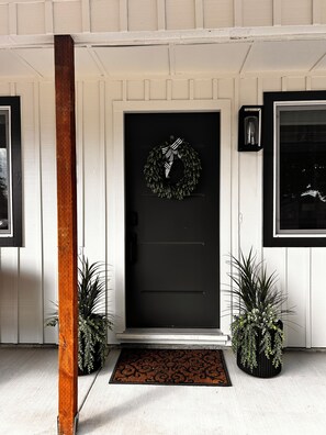 A black front door with a wreath, a doormat, and two potted plants on either side.