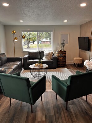 A modern living room with a leather sofa, green armchairs, a round coffee table, and a large window with a view of trees and a building.