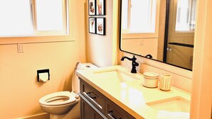 Et badeværelse med toilet, håndvask og spejl. Væggene er dekoreret med billeder, og der er et vindue over toilettet.