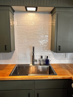 A kitchen with a stainless steel sink, a faucet, and a backsplash made of white tiles. There are two bottles on the counter, and a light fixture above the sink.
