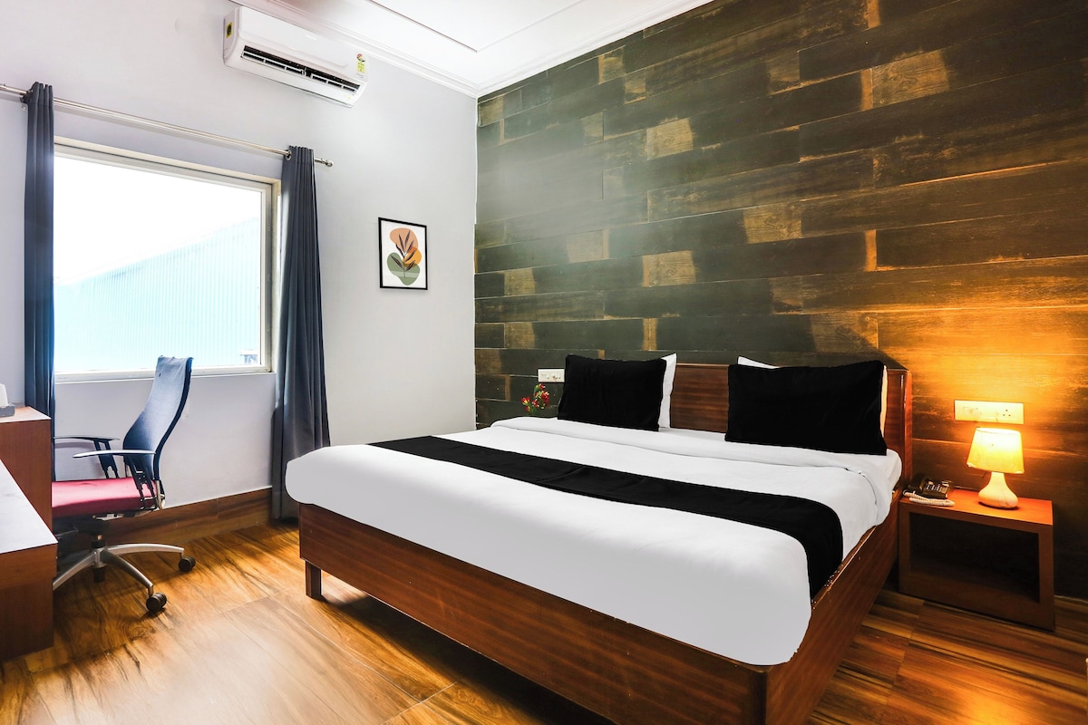 Una habitación de hotel con suelo de madera, cama con ropa de cama blanco y negro, un escritorio con silla, ventana con cortinas y aire acondicionado instalado en la pared.