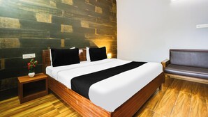Una habitación de hotel con piso de madera, cama con ropa blanca, mesita de noche con un florero y un sofá.