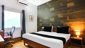 Una habitación de hotel con suelo de madera, cama con ropa de cama blanco y negro, un escritorio con silla, ventana con cortinas y aire acondicionado instalado en la pared.