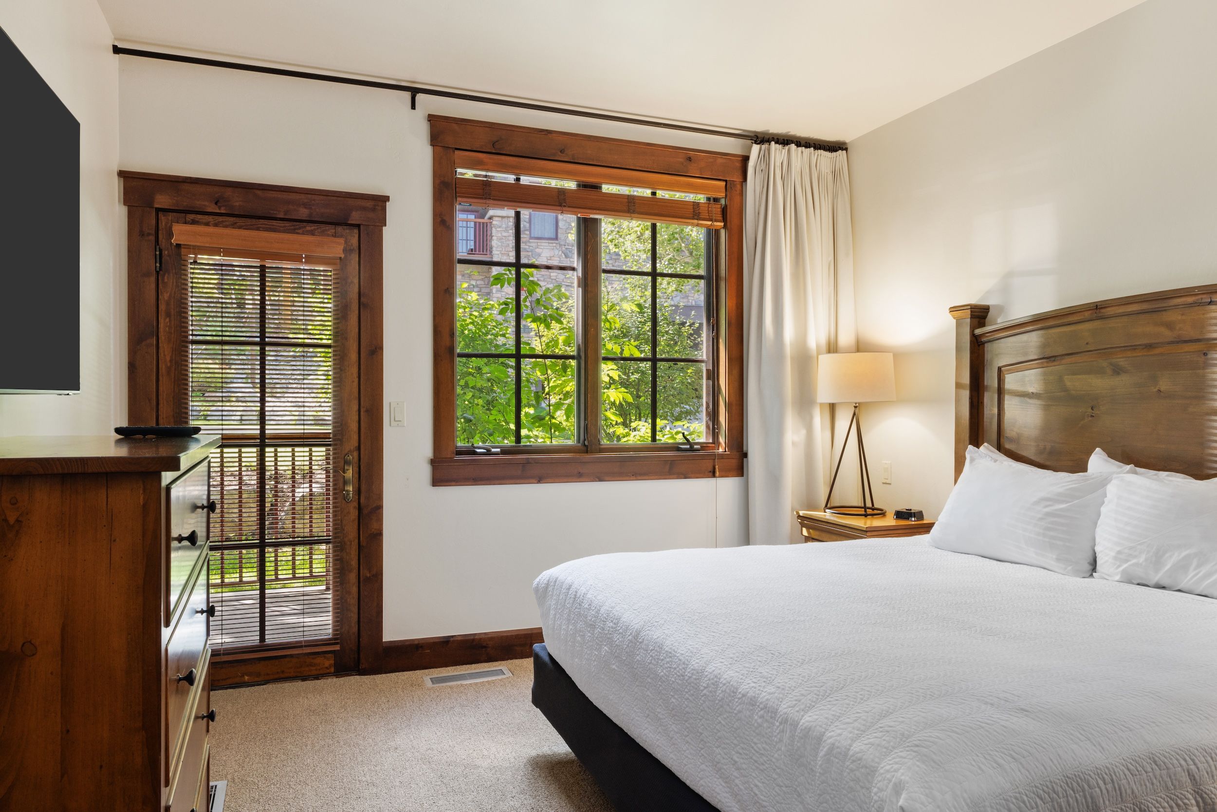 A bedroom with a bed, a nightstand, a lamp, a window with a view of greenery, and a door with blinds.