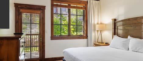 A bedroom with a bed, a nightstand, a lamp, a window with a view of greenery, and a door with blinds.