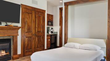 A bedroom with a bed, a television mounted on the wall, a fireplace, wooden wardrobes, and a kitchen area with a microwave.