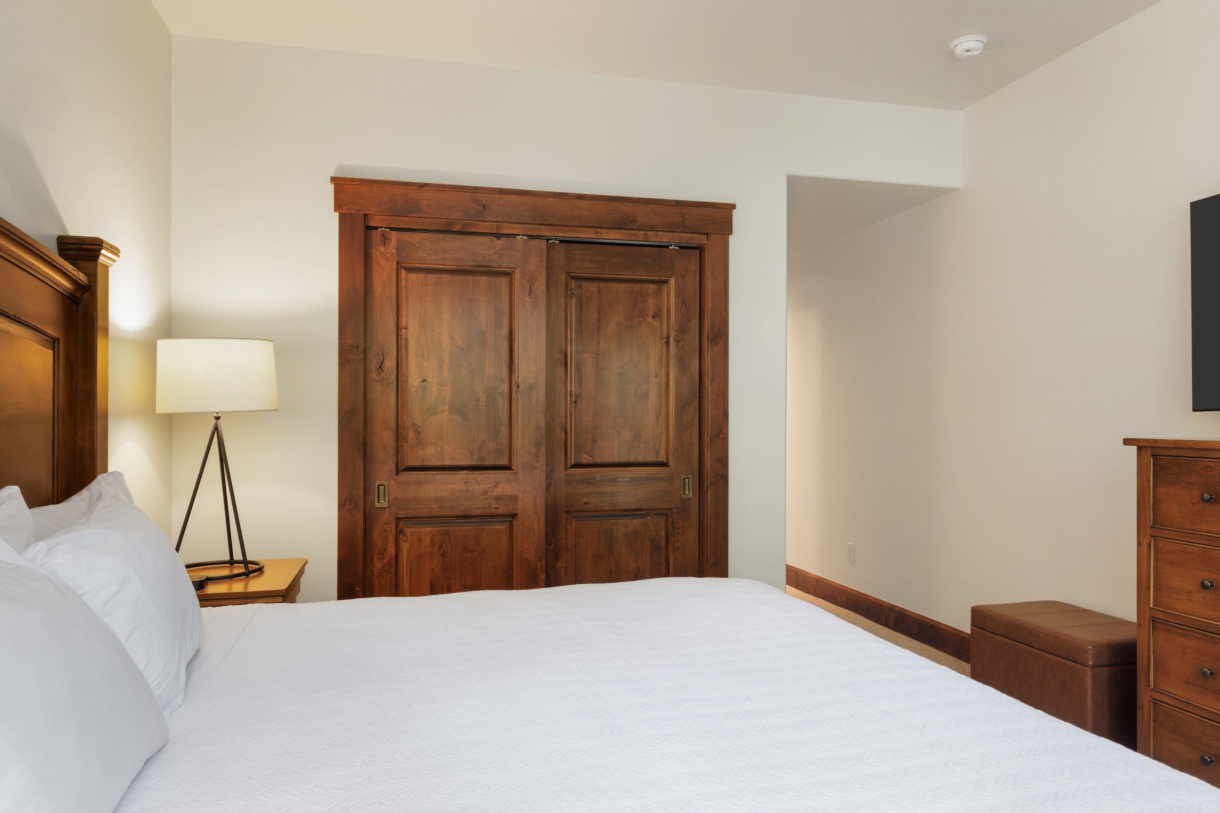 A bedroom with a large bed, wooden wardrobe, bedside table with lamp, and a dresser.