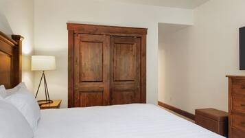 A bedroom with a large bed, wooden wardrobe, bedside table with lamp, and a dresser.