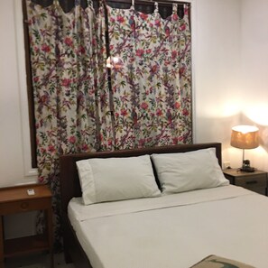 Ein Bett mit weißer Bettwäsche, ein Nachttisch mit einer Lampe und ein Fenster mit floralen Vorhängen.