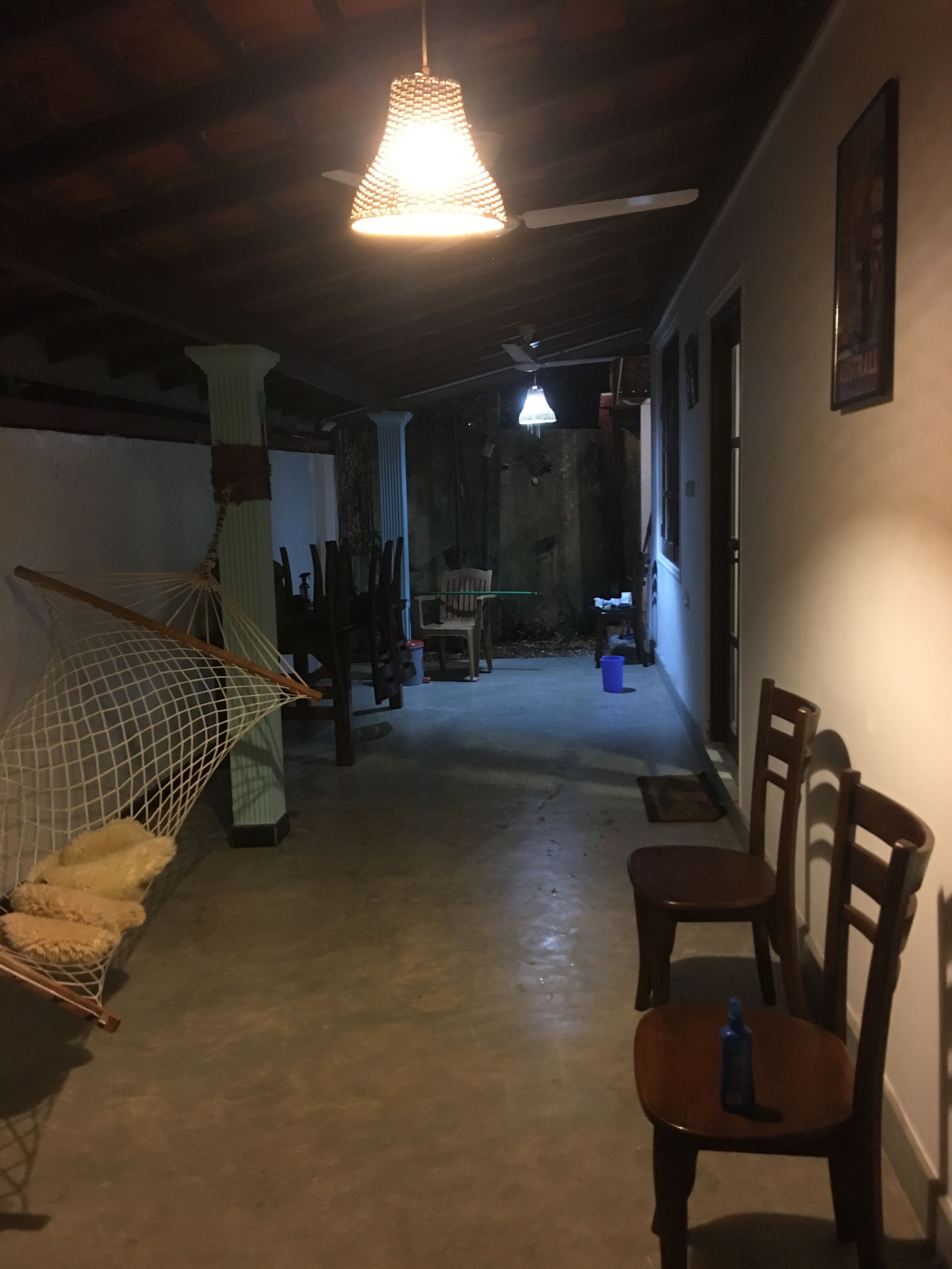 Un couloir faiblement éclairé, avec un hamac, des chaises et un ventilateur de plafond.