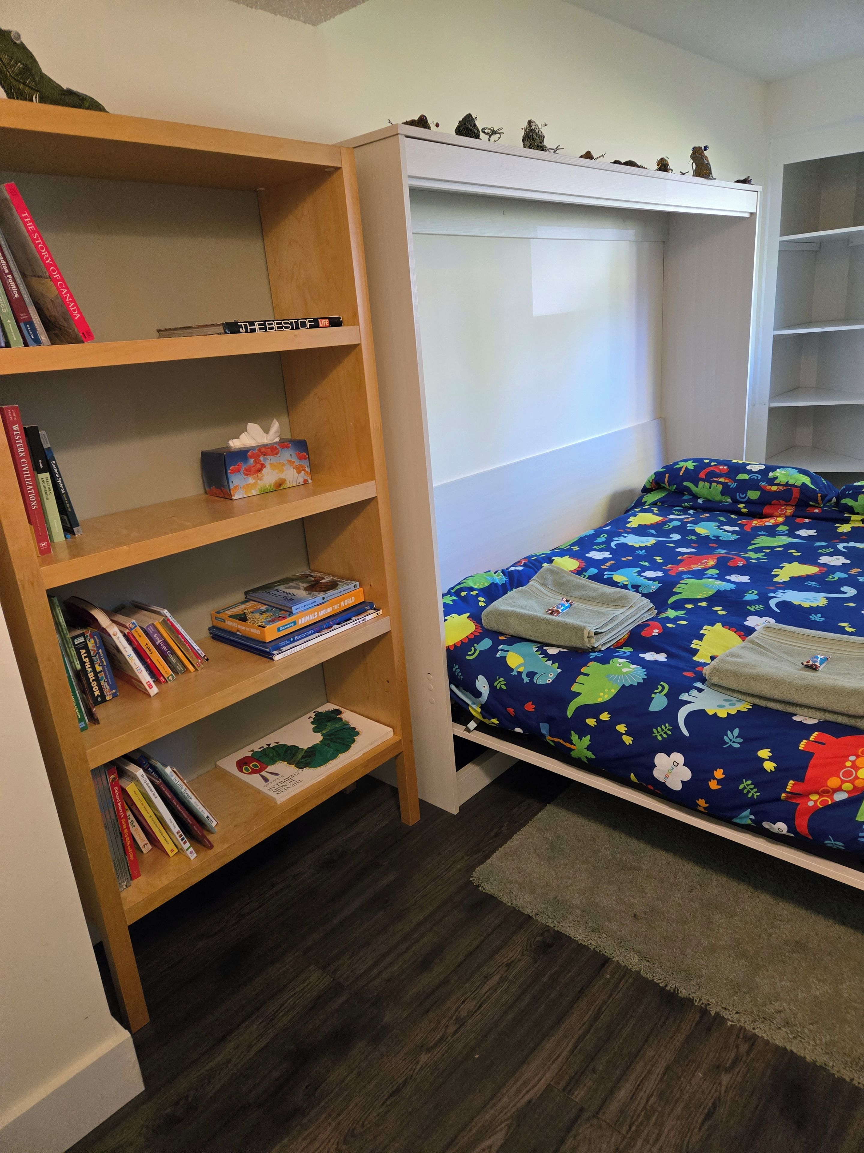 A bedroom with a wooden bookshelf, a bed with a colorful dinosaur-themed bedspread, and a white headboard.