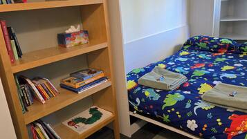 A bedroom with a wooden bookshelf, a bed with a colorful dinosaur-themed bedspread, and a white headboard.