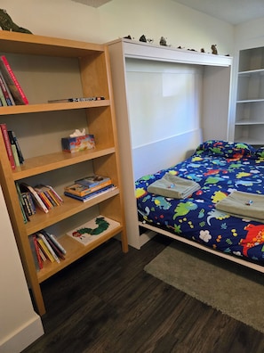 A bedroom with a wooden bookshelf, a bed with a colorful dinosaur-themed bedspread, and a white headboard.