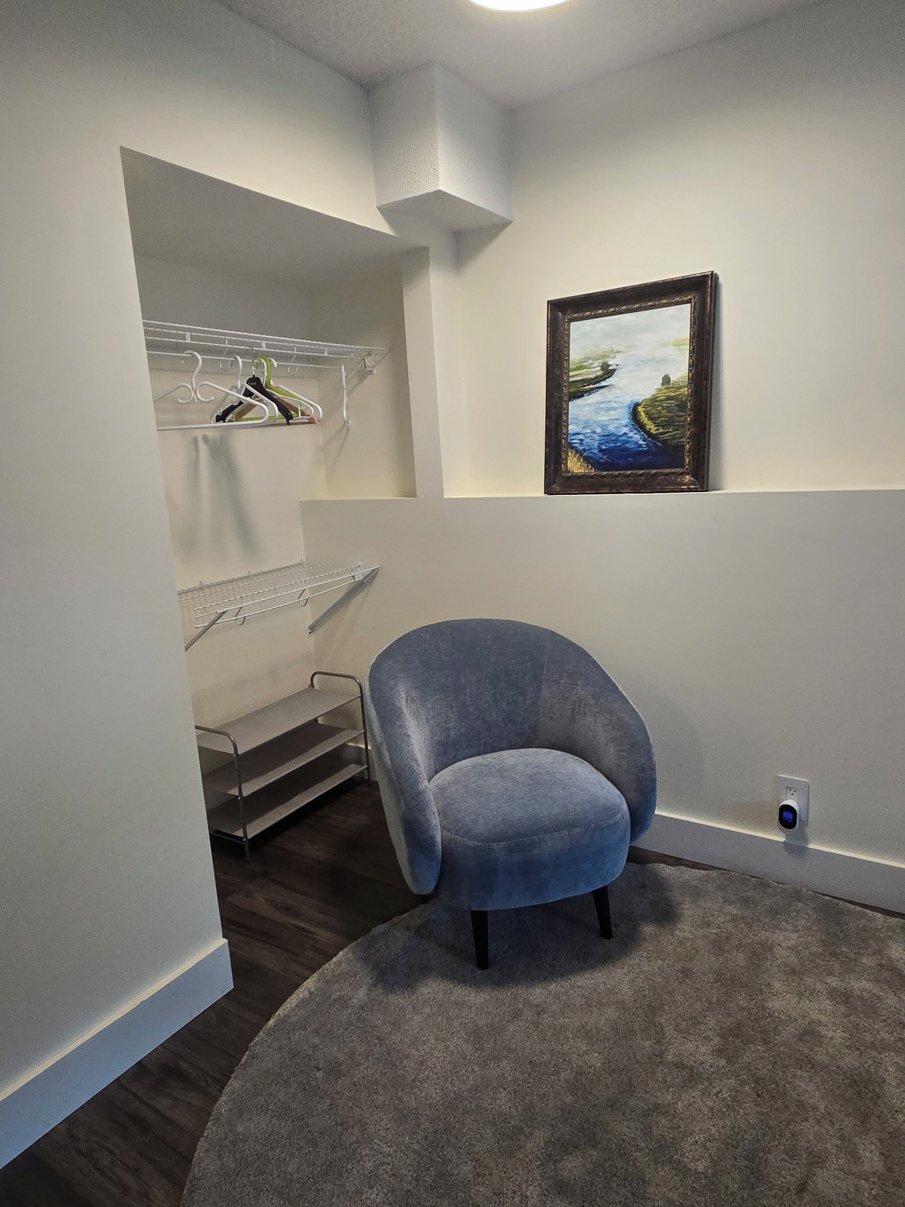 A room with a grey armchair, a painting on the wall, a white shelving unit, and a grey carpet.