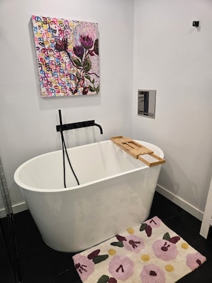 A modern bathroom with a freestanding bathtub, a wooden tray, a colorful abstract painting, and a fruit-patterned rug. - Luxury Family and Pet Friendly Apartment (Calgary)