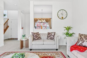 A living room with a sofa, armchairs, a clock, and a window with curtains.