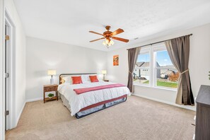 A bedroom with a bed, a nightstand, a ceiling fan, a window with curtains, and a view of the outside.