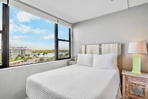 A hotel room with a large window, a bed with white bedding, a nightstand with a lamp, and a view of a body of water and buildings. - Harbor Towers 515 in Siesta Key (Siesta Key)