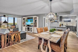 A modern living room with a large TV, a dining area with wicker chairs, and a kitchen with a stone backsplash. - Harbor Towers 515 in Siesta Key (Siesta Key)