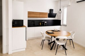 Een compacte keuken met een eethoek, met een zwarte afzuigkap, witte kastjes en een ronde houten tafel met witte stoelen.