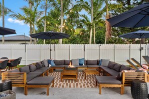 An outdoor seating area with a wooden sectional sofa, a fire pit, and multiple umbrellas. - Saltwater Pool -12Hole Miniature Golf & Pickleball (South Miami Heights)