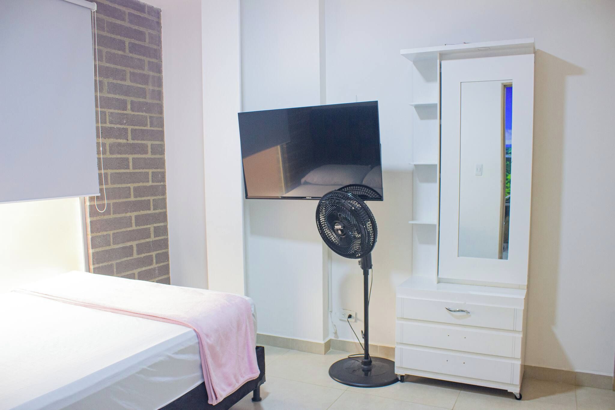 Un dormitorio con una cama, un televisor montado en la pared, un ventilador de pie y un armario.