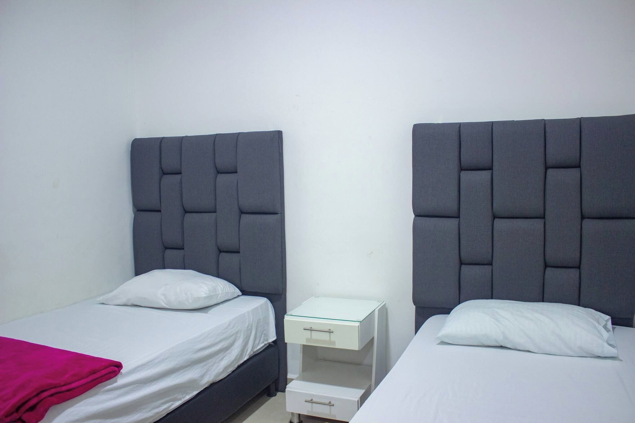 A room with two single beds, each with a pillow and a white blanket. There is a small bedside table with a drawer and a glass top.
