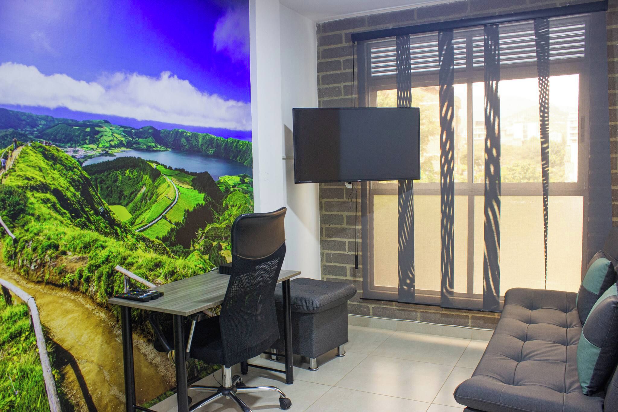 Una habitación con escritorio, silla de oficina y sofá. Hay un televisor montado en la pared y una ventana con persianas.