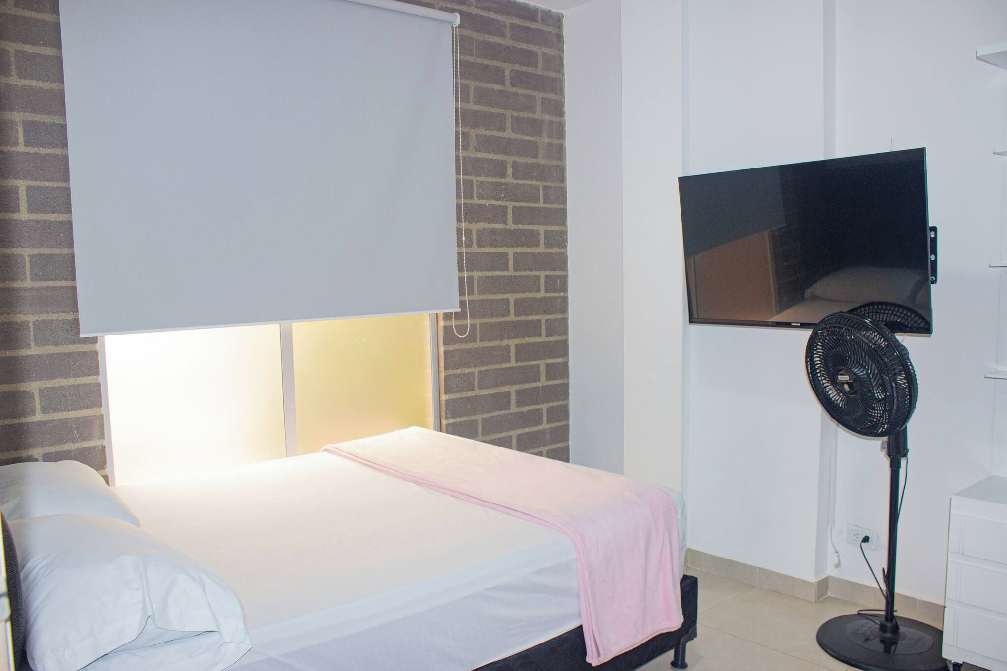 Un dormitorio con una cama, un televisor montado en la pared y un ventilador de pie.