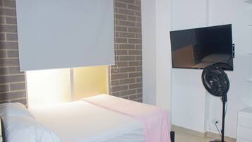 Un dormitorio con una cama, un televisor montado en la pared y un ventilador de pie.
