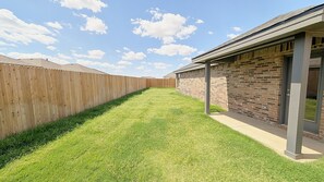 A backyard with a wooden fence, a grassy lawn, and a brick building with a covered patio area. - Modern Midland Retreat (Midland)