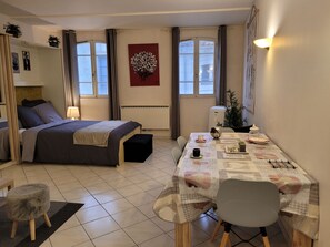 Ein gemütliches Schlafzimmer mit Bett, Nachttisch und einem kleinen Tisch mit Stuhl. Es gibt ein Fenster mit Vorhängen, eine Heizung und eine Wandleuchte.