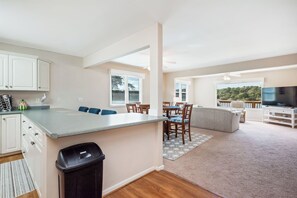 Cocina moderna con una isla grande, comedor con sillas, sala de estar con sofá y televisor, y vista a zonas verdes al exterior.